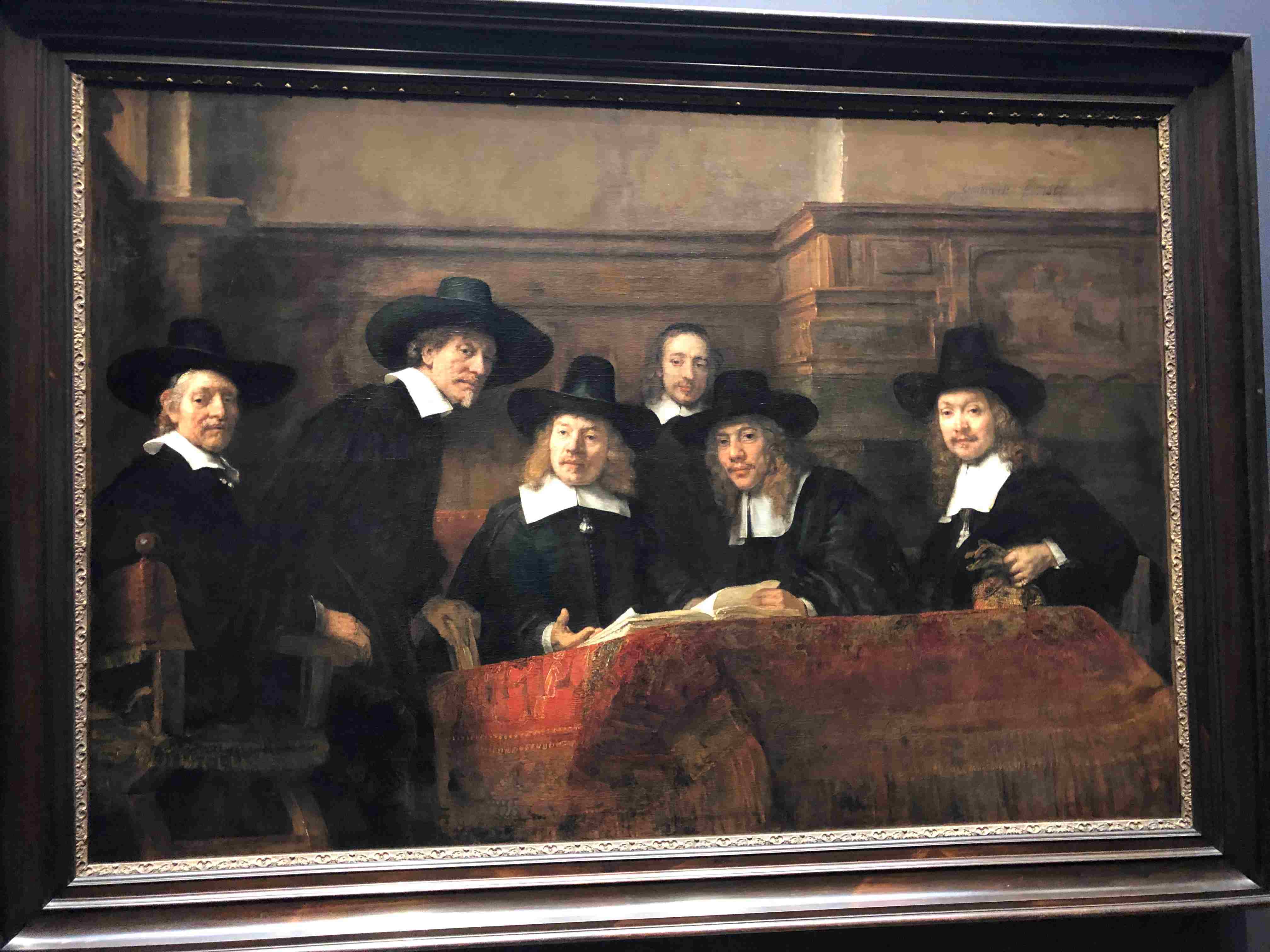 Famous Dutch Masters painting with six gentlemen with black hats, white wide collars, black suits turning the pages of a book together